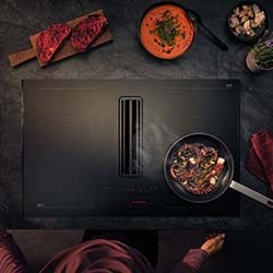 A young girl in a brown dress with black dots rolls dough on a wooden table beside a black stand mixer and a jar of flour. A cinnamon roll sits nearby.