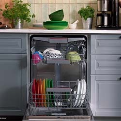 Open dishwasher in a modern kitchen with colorful dishes and bowls neatly organized inside. Fresh herbs and a coffee maker sit on the countertop above.