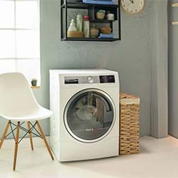 Modern laundry room with a front-loading washing machine, white chair, and wicker laundry basket. Shelves with towels and decor create a tidy, serene feel.