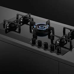 Modern black glass gas stovetop with five burners, blue flame lit on one, and five control knobs. Sleek, minimalist design on a dark countertop.