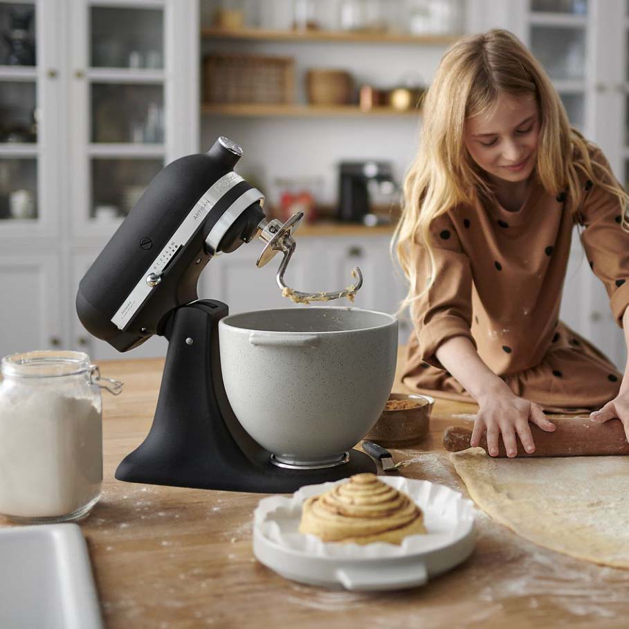 A young girl in a brown dress with black dots rolls dough on a wooden table beside a black stand mixer and a jar of flour. A cinnamon roll sits nearby.