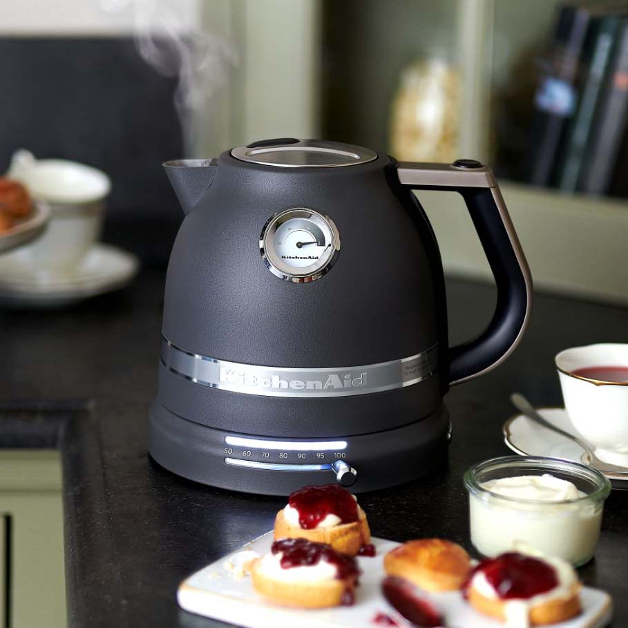Black electric kettle emitting steam on a kitchen counter, surrounded by scones with jam and cream.