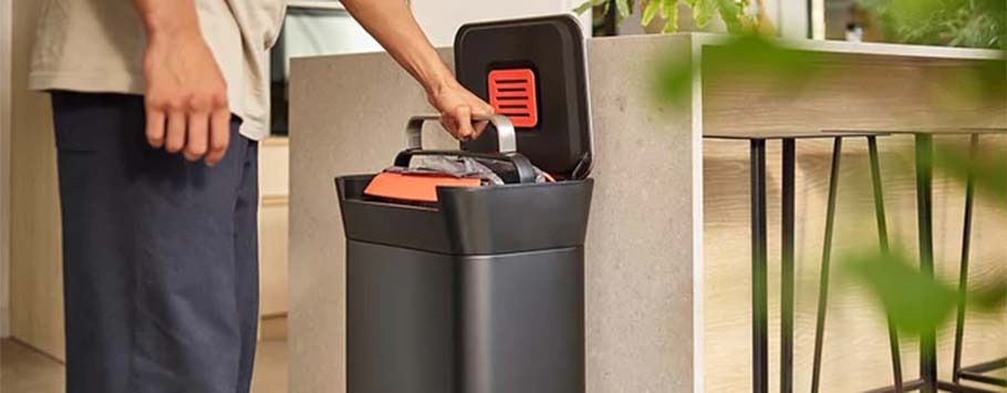 A person in casual attire uses a black trash compactor in a modern kitchen. The compactor is open, and green plants frame the scene, creating a tidy, eco-friendly setting.