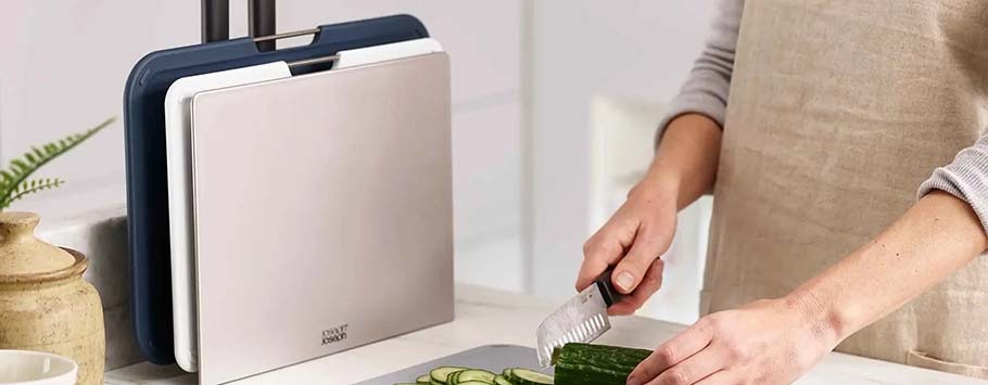 A person in an apron slices a cucumber on a black cutting board. Nearby, a set of color-coded cutting boards is stored upright, creating a tidy kitchen scene.