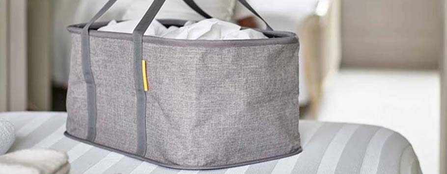 A gray fabric storage basket with handles sits on a striped bedspread, filled with white towels. The scene conveys a tidy, calm bedroom setting.