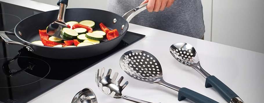 A person cooks sliced zucchini and red bell peppers in a black frying pan on an induction cooktop. Four stainless steel kitchen utensils rest on a white countertop.
