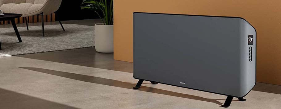Modern gray heater with digital display placed on a sleek, polished floor in a cosy living room. A beige armchair and a potted plant are in the background.