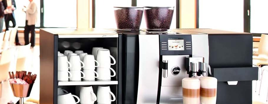 A sleek coffee machine with touchscreen in a bright room. Coffee cups are neatly arranged nearby, and two lattes in glass cups sit in front.
