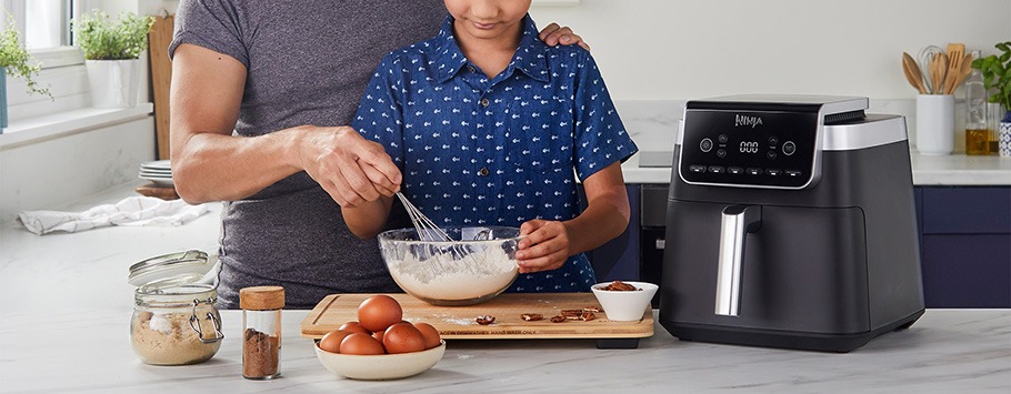 A parent and child mix batter in a bowl beside an air fryer on a kitchen counter. Ingredients like eggs and sugar surround them. The scene is warm and collaborative.