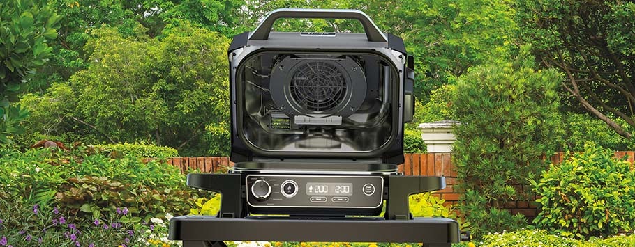 A sleek, black air fryer stands open on a patio, surrounded by lush greenery. The digital display shows settings, evoking a modern, outdoor cooking vibe.