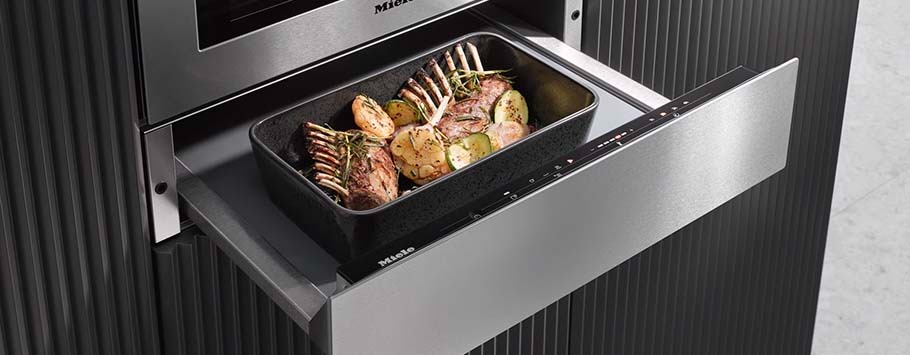 A sleek oven with an open warming drawer containing a roasting pan filled with seasoned lamb racks and sliced potatoes, exuding warmth and culinary precision.