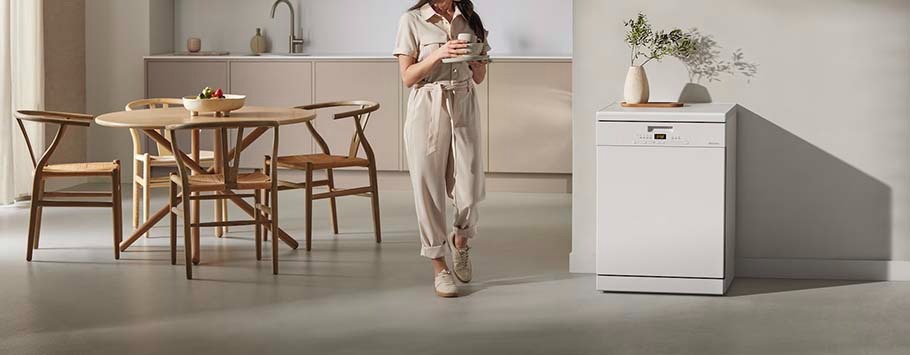 A woman in a beige jumpsuit holds a mug, walking in a modern kitchen with a round wooden table, chairs, and a white dishwasher. The ambiance is calm and minimalist.
