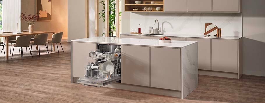 Modern kitchen with beige cabinets, featuring a central island with an open dishwasher loaded with dishes. Warm lighting and wood flooring add a cosy feel.