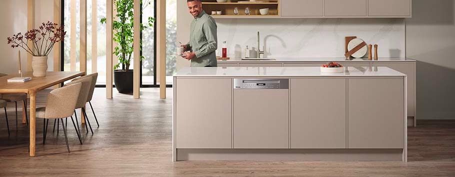 Modern kitchen with a man smiling, holding a pot. Beige cabinets, white countertop, wooden dining table, and indoor plants. Bright and minimalist ambiance.