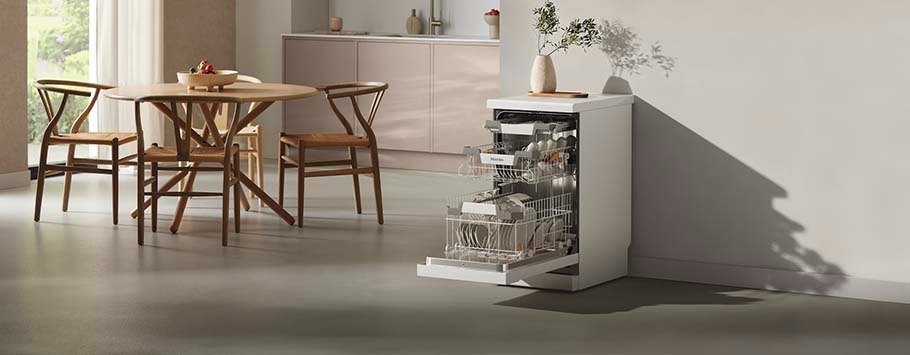 Modern kitchen with an open, fully loaded dishwasher on the right, a round wooden dining table with four chairs on the left, and sunlight streaming in.