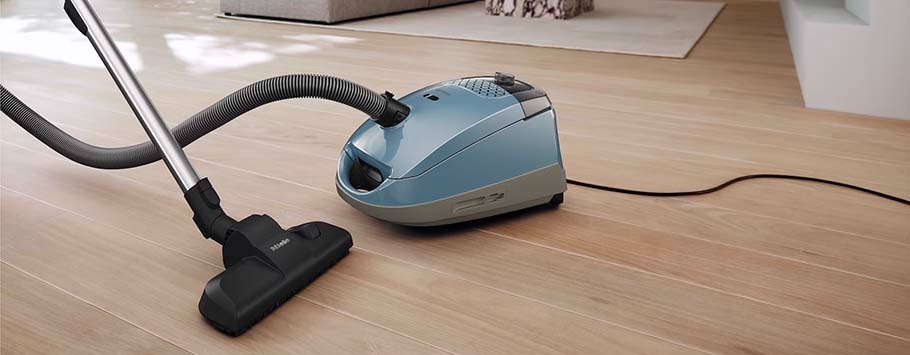 A light blue canister vacuum cleaner is on a wooden floor. The hose and nozzle are visible, suggesting cleaning action. Sunlight shines softly on the floor, creating a tidy, peaceful atmosphere.