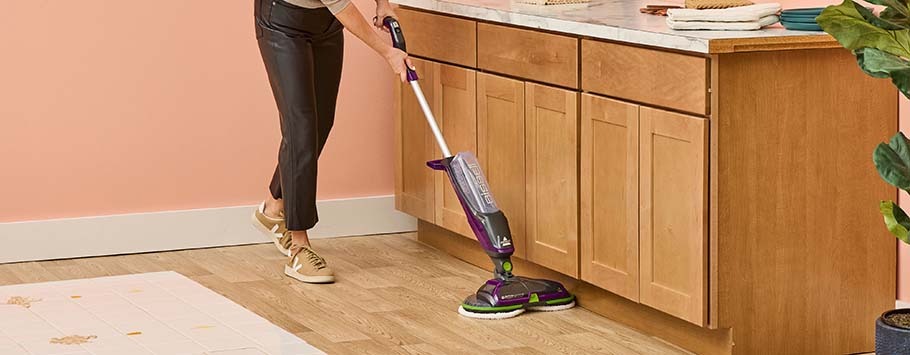 A person cleans a wooden kitchen floor with a purple and white motorized mop. The space is bright and tidy, evoking a sense of cleanliness and efficiency.