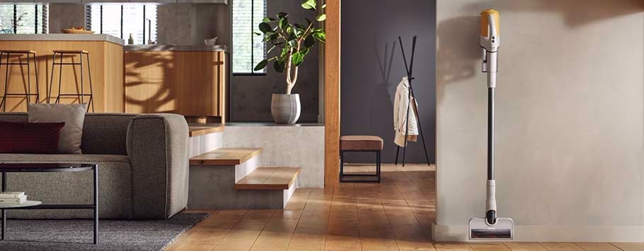 Modern living room with a sleek cordless vacuum mounted on a wall. Features wooden floors, a gray sofa, and bar stools, exuding a clean and cosy feel.