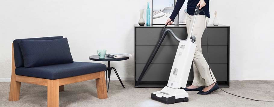 A person vacuuming a light grey carpet in a modern room, featuring a dark chair and table. The setting is clean and minimalistic, conveying a calm tone.