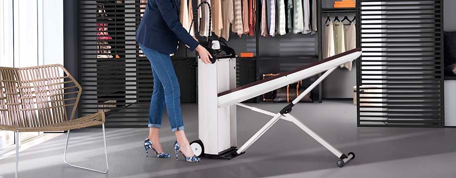 A woman adjusts a collapsible ironing board in a modern, organized laundry room with hanging clothes. The setting is bright and stylish, conveying efficiency.