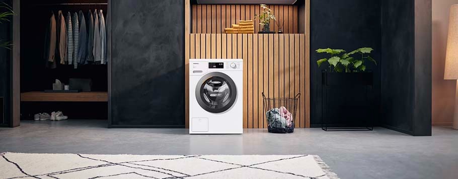 Modern laundry room with a sleek white washer centred against a wood accent wall. Nearby are folded towels, a plant, and a basket with clothes, conveying a tidy, minimalist vibe.