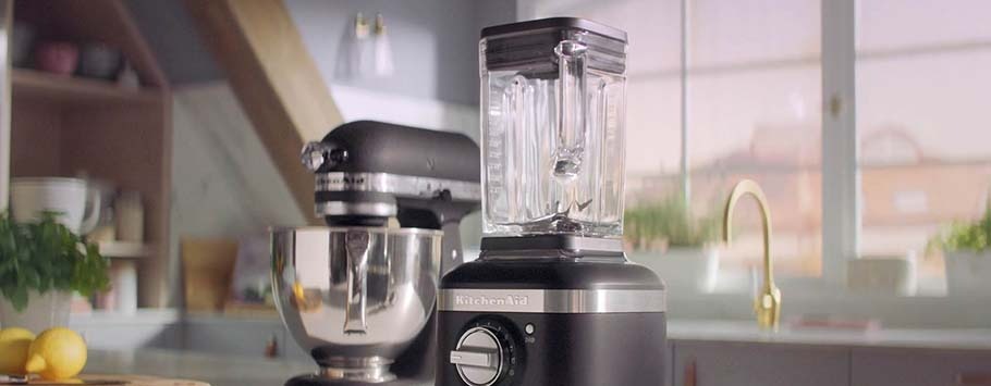 A sleek black KitchenAid blender sits on a modern kitchen counter, next to a black stand mixer. Sunlight streams through a window, creating a warm, inviting atmosphere.