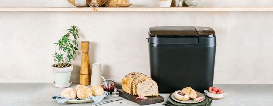 Black compost bin on a kitchen counter with freshly baked bread, rolls, a potted plant, and a pepper mill. The setting is neat and inviting.
