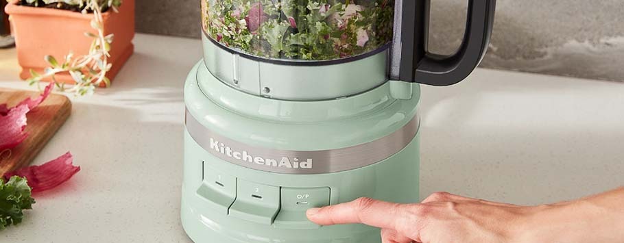A person’s hand is about to press the start button on a pastel green KitchenAid food processor filled with chopped greens. A cutting board with leaves and a potted plant are in the background. The scene conveys a fresh, culinary preparation theme.