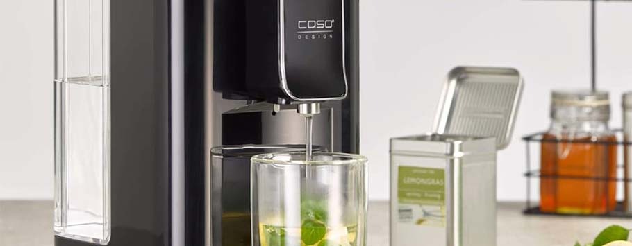 A sleek black appliance dispensing hot water into a glass with herbs. Nearby, a lemongrass tea tin and honey jar suggest a coey tea-making scene.
