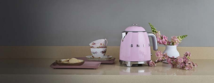 A pink kettle on a beige countertop with a floral teacup set, almond cookies on a pink tray, and delicate pink flowers in a white vase, creating a cosy, elegant ambiance.