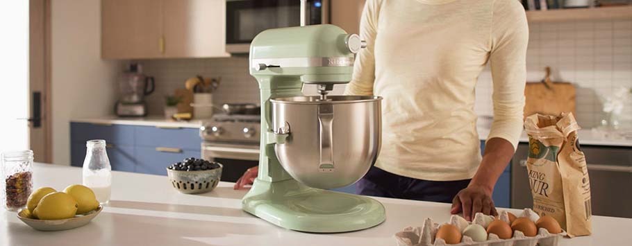 A person stands in a kitchen beside a pale green stand mixer. Nearby are eggs, flour, and lemons, with a modern stove and cabinets in the background.