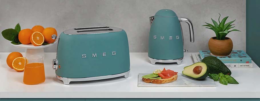 Retro-style teal toaster and kettle on a countertop with a plate of avocado toast, sliced avocado, oranges, a glass of juice, and a potted plant nearby.