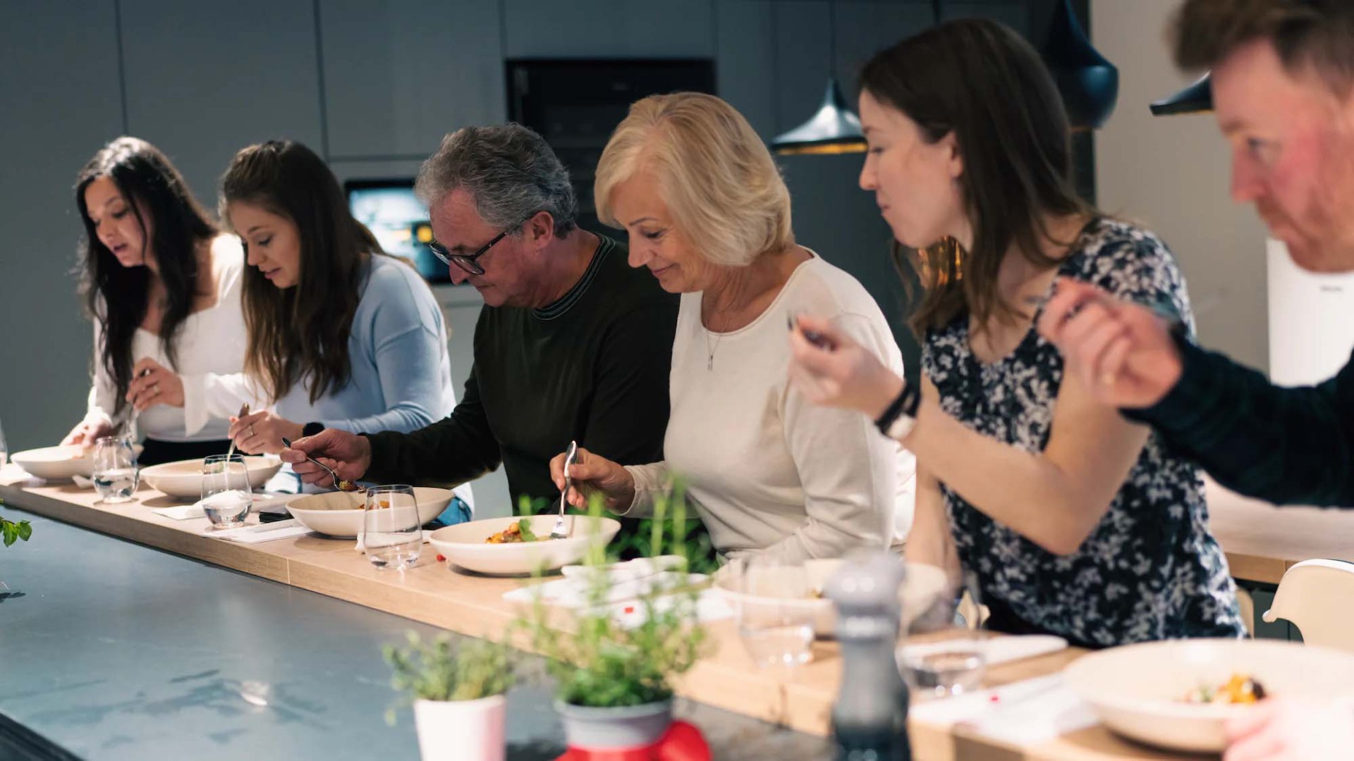 A group of six people sit at a bar in a modern kitchen, tasting food from white bowls. They appear focused and engaged.