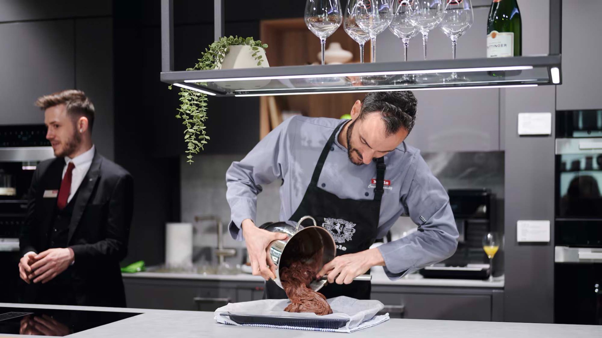 Chef pouring chocolate mix onto a tray in a modern kitchen. A man in a suit observes. Wine glasses and a plant hang above.