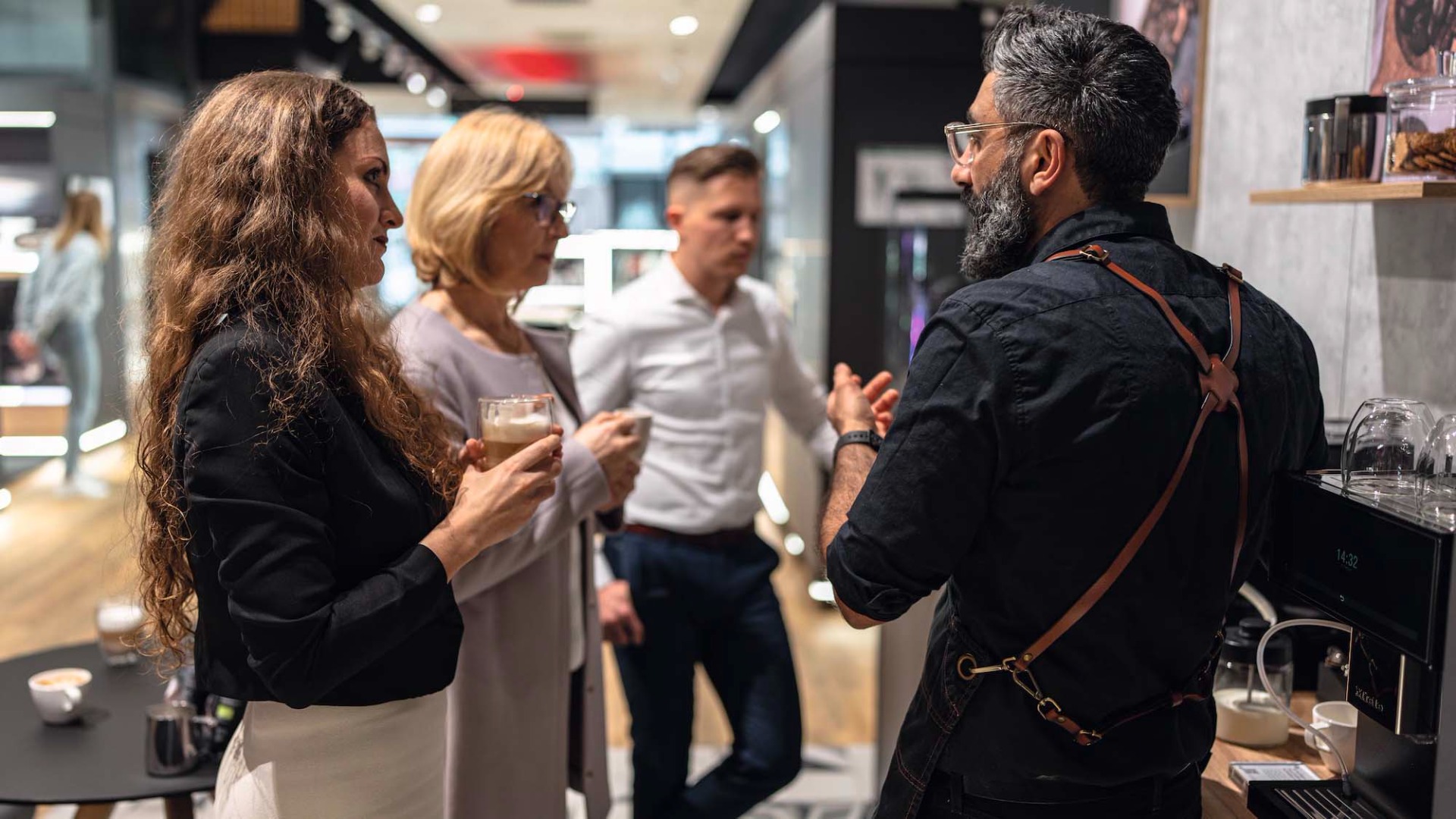 A bearded man in an apron explains coffee-making to three attentive adults in a stylish cafe.