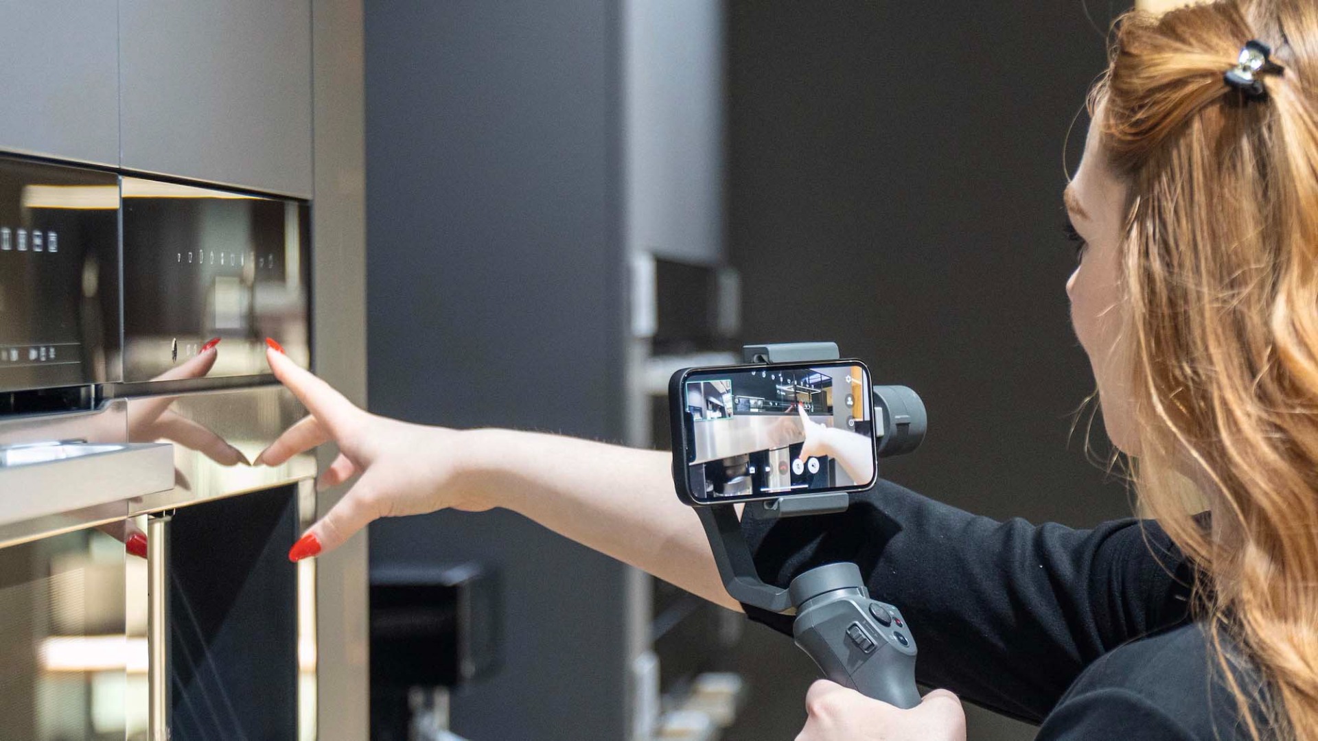 A woman films a modern appliance with her smartphone mounted on a stabilizer. Her red nails and black attire contrast with the sleek, metallic finish.