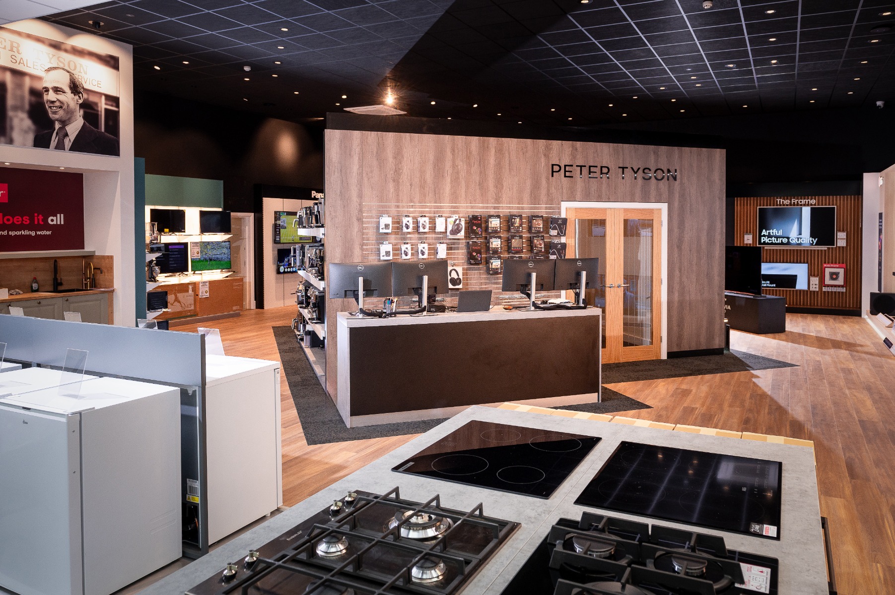 Modern electronics store interior with wood panelling, computer displays, and sleek kitchen appliances. Bright lighting and organized layout.